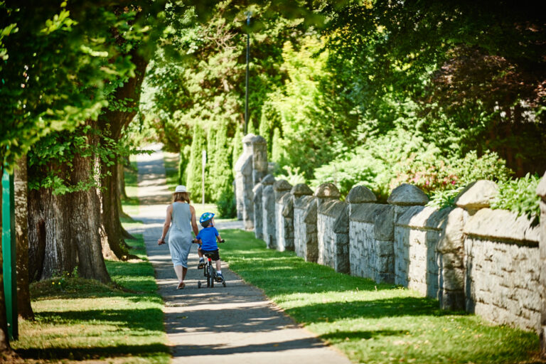 Parent walking beside a child riding a bicycle along a tree-lined sidewalk in Vancouver’s Oakridge neighbourhood.